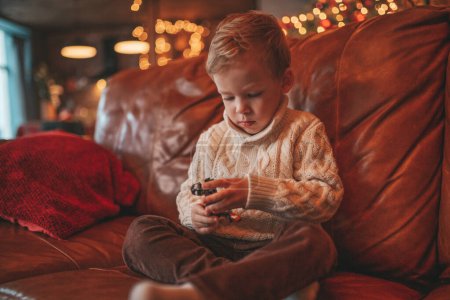 Photo for Smile small child being active having fun in motion and dreamer waiting for miracle Santa. Cheerful kid in knitwear outfit playing with toy indulge indoor on sofa eve 25 December garlands lights Noel - Royalty Free Image