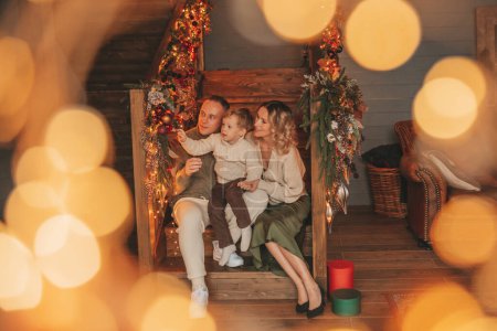 Photo for Portrait of happy family in knitted beige sweaters waiting Santa indoor. Smiling parents hugs and kisses his little son eve 25 December celebrates xmas with garlands lights Noel tree gifts giving - Royalty Free Image