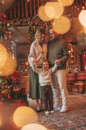 Photo for Portrait of happy family in knitted beige sweaters waiting Santa indoor. Smiling parents hugs and kisses his little son eve 25 December celebrates xmas with garlands lights Noel tree gifts giving - Royalty Free Image