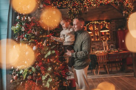 Photo for Smile little boy and dad having fun with each other hugs and kisses indoor waiting santa. Celebrating New year with garlands lights of Noel tree winter season Xmas spirit eve 25 December paternal love - Royalty Free Image