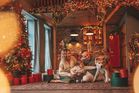 Photo for Portrait of happy family in knitted beige sweaters waiting Santa indoor with Akita Inu. Smiling parents hugs and kisses his little son eve 25 December celebrates xmas with garlands lights Noel tree - Royalty Free Image