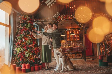 Photo for Portrait of happy family in knitted beige sweaters waiting Santa indoor with Akita Inu. Smiling parents hugs and kisses his little son eve 25 December celebrates xmas with garlands lights Noel tree - Royalty Free Image