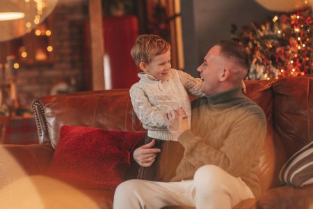 Photo for Smile little boy and dad having fun with each other hugs and kisses indoor waiting santa. Celebrating New year with garlands lights of Noel tree winter season Xmas spirit eve 25 December paternal love - Royalty Free Image