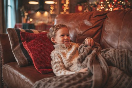 Photo for Smile small child being active having fun in motion and dreamer waiting for miracle Santa. Cheerful kid in knitwear outfit playing with toy indulge indoor on sofa eve 25 December garlands lights Noel - Royalty Free Image