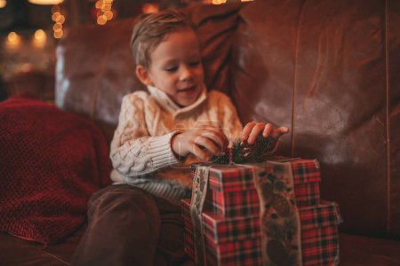 Photo for Smile small child being active having fun waiting for miracle Santa open presents. Cheerful kid in casual knit outfit celebrating new year with gifts boxes garlands bokeh lights noel eve 25 december - Royalty Free Image