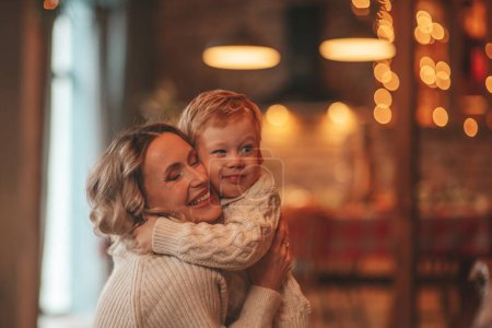 Photo for Portrait of happy family mom with small son in knitted beige sweaters waiting Santa indoor. Mothers love hugs and kisses in eve 25 December celebrate New year gifts giving garlands lights Noel tree - Royalty Free Image