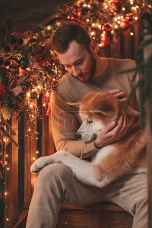 Portrait of young lovely couple hugging indoor eve 25 December with akita inu dog. People in knit stylish outfit hugs kisses tenderness celebrating new year garlands xmas lights noel tree at home