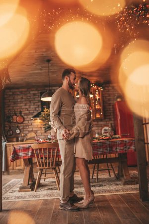 Photo for Portrait of young lovely couple hugging indoor eve 25 December. Lovers laughing hugs kisses waiting xmas at home. Celebrating new year garlands lights noel in elegant knitwear outfit tenderness - Royalty Free Image