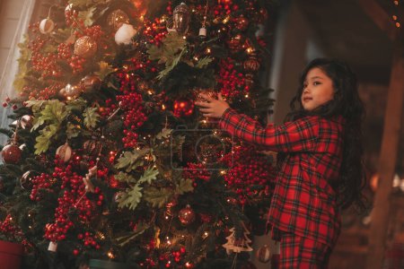 Happy cute japan little girl dreamer waiting miracle from Santa posing decorates noel tree. Asian kid black curly hair in red plaid outfit celebrating new year at bokeh xmas lights eve 25 december
