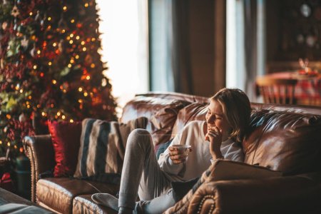Young emotional cheerful teen boy with long hair laughter and joyful at cozy home. Stylish zoomer gen Z in good mood celebrates new year holidays with xmas tree bokeh lights garlands eve 25 december