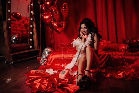 Foto de Sexy modelo sensual asiática en ropa interior de encaje rojo y boa de plumas posando con teléfono vintage rosa cerca de la cama en el día de San Valentín - Imagen libre de derechos
