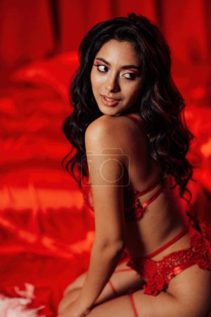 Photo for Side view of curly black haired asian sensual model seduces sitting on the kneel on the floor at red glamour background - Royalty Free Image