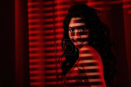 Foto de Rizado negro pelo asiático sensual modelo bajo luz y sombra de persianas en la piel en rojo glamour fondo. - Imagen libre de derechos
