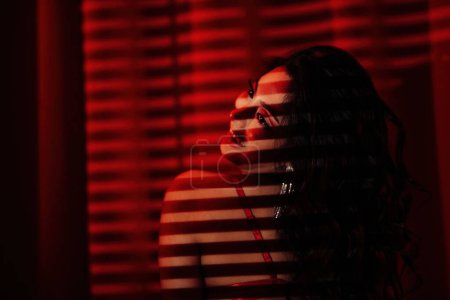 Foto de Curly black hair asian sensual model under light and shadow of blinds on skin at red glamour background. - Imagen libre de derechos