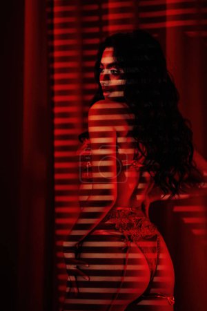 Foto de Joyful hot asian model in lace underwear under light and shadow of blinds at red glamour background - Imagen libre de derechos