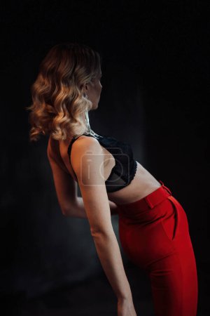 Téléchargez les photos : Young adult beauty woman in formal evening suit of red color with lace black bra underwear standing by thoughtful. Stylish blonde curly hair model fashionista posing at studio in fashion pantsuit - en image libre de droit