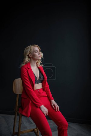 Photo for Young adult beauty woman in formal evening suit of red color with lace black bra underwear standing by thoughtful. Stylish blonde curly hair model fashionista posing at studio in fashion pantsuit - Royalty Free Image