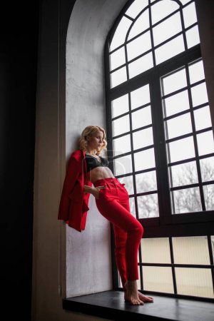 Foto de Young adult beauty woman in formal evening suit of red color with lace black bra underwear standing by thoughtful. Stylish blonde curly hair model fashionista posing at studio in fashion pantsuit - Imagen libre de derechos