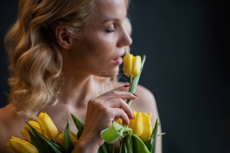 Foto de Adult beauty woman half naked in formal evening red trousers without bra hugs bouquet of yellow tulips. Stylish blonde curly hair sensual nude model fashionista posing in studio at spring holidays - Imagen libre de derechos