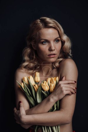 Téléchargez les photos : Adult beauty woman half naked in formal evening red trousers without bra hugs bouquet of yellow tulips. Stylish blonde curly hair sensual nude model fashionista posing in studio at spring holidays - en image libre de droit
