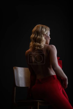 Adult beauty woman bare back in formal evening red trousers sitting pose without bra. Stylish blonde curly hair sensual nude model fashionista posing at studio in fashion pantsuit out of blazer