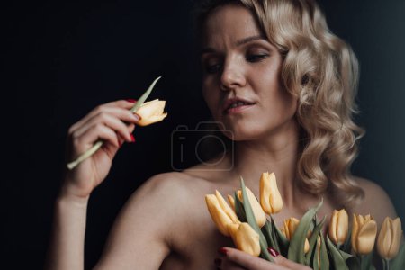 Photo for Adult beauty woman half naked in formal evening red trousers without bra hugs bouquet of yellow tulips. Stylish blonde curly hair sensual nude model fashionista posing in studio at spring holidays - Royalty Free Image
