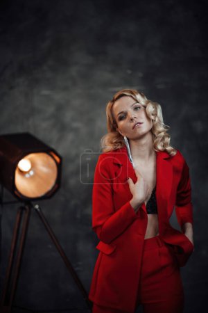 Téléchargez les photos : Jeune femme de beauté adulte en costume de soirée formelle de couleur rouge avec dentelle soutien-gorge noir sous-vêtements debout par réfléchi. Élégant blonde cheveux bouclés mannequin fashionista posant en studio en costume de mode - en image libre de droit