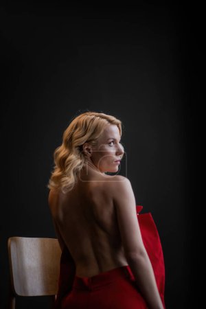 Téléchargez les photos : Adult beauty woman bare back in formal evening red trousers sitting pose without bra. Stylish blonde curly hair sensual nude model fashionista posing at studio in fashion pantsuit out of blazer - en image libre de droit