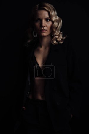 Téléchargez les photos : Young adult beauty woman in formal evening suit of black color with lace bra at thoughtful. Stylish blonde curly hair sensual model fashionista posing at studio in fashion pantsuit - en image libre de droit