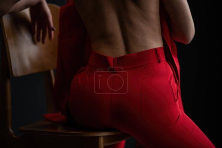 Téléchargez les photos : Adult beauty woman bare back in formal evening red trousers sitting pose without bra. Stylish blonde curly hair sensual nude model fashionista posing at studio in fashion pantsuit out of blazer - en image libre de droit