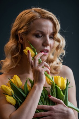 Photo for Adult beauty woman half naked in formal evening red trousers without bra hugs bouquet of yellow tulips. Stylish blonde curly hair sensual nude model fashionista posing in studio at spring holidays - Royalty Free Image