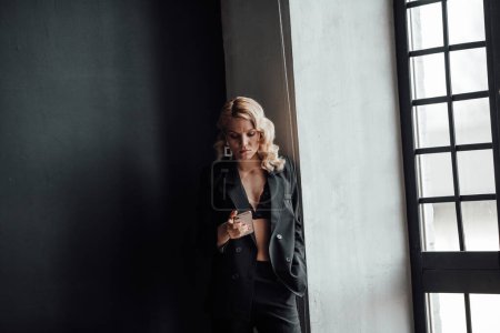 Foto de Adult beauty woman in formal evening suit of black color with lace bra taking photo for social media. Stylish blonde curly hair model fashionista posing in fashion pantsuit no shirt video calling - Imagen libre de derechos