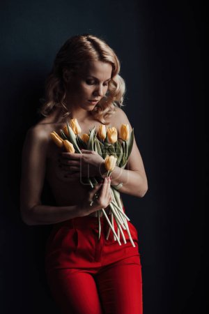 Foto de Adult beauty woman half naked in formal evening red trousers without bra hugs bouquet of yellow tulips. Stylish blonde curly hair sensual nude model fashionista posing in studio at spring holidays - Imagen libre de derechos