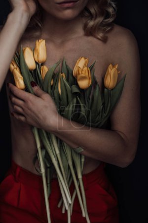 Foto de Mujer de belleza adulta medio desnuda en pantalones rojos formales de noche sin sujetador abraza ramo de tulipanes amarillos. Elegante rubia rizado pelo sensual modelo desnudo fashionista posando en el estudio en las vacaciones de primavera - Imagen libre de derechos