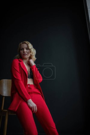 Foto de Young adult beauty woman in formal evening suit of red color with lace black bra underwear standing by thoughtful. Stylish blonde curly hair model fashionista posing at studio in fashion pantsuit - Imagen libre de derechos