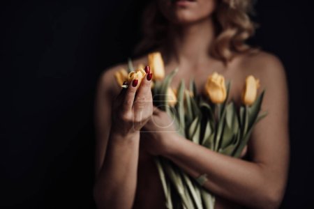 Foto de Mujer de belleza adulta medio desnuda en pantalones rojos formales de noche sin sujetador abraza ramo de tulipanes amarillos. Elegante rubia rizado pelo sensual modelo desnudo fashionista posando en el estudio en las vacaciones de primavera - Imagen libre de derechos