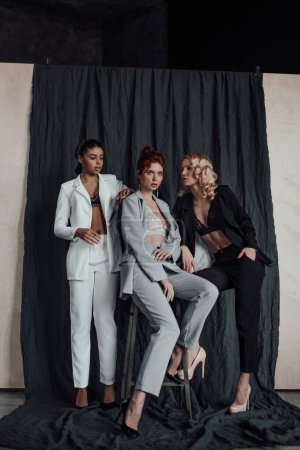 Photo for Adult beauty elegant multi ethnic group young girls in formal evening suits of different colors with bra. Stylish diverse people sensual fashionistas posing at studio in fashion pantsuits no shirts - Royalty Free Image