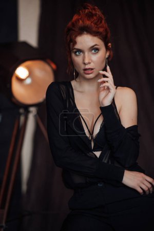 Photo for Adult beauty elegant young woman in formal evening black clothing hand on face touching. Stylish ginger curly hair sensual model bare shoulder fashionista posing at studio in fashion pantsuit and bra - Royalty Free Image