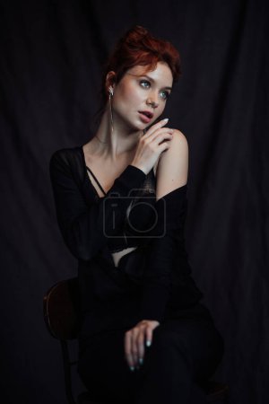 Photo for Adult beauty elegant young woman in formal evening black clothing hand on face touching. Stylish ginger curly hair sensual model bare shoulder fashionista posing at studio in fashion pantsuit and bra - Royalty Free Image