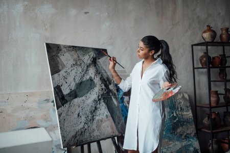 Young artist beauty swarthy woman in white long shirt bare legs posing against her paintings. Stylish black curly hair sensual african american model with paint palette and brushes at painting studio