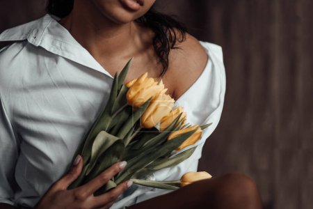 Jeune femme adulte beauté swarthy en chemise blanche épaules nues étreintes bouquet de tulipes jaunes. élégant noir bouclé cheveux sensuel afro-américain modèle fashionista posant en studio pendant les vacances de printemps