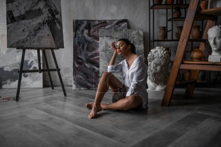 Photo for Young adult beauty swarthy woman in white shirt bare shoulder hugs ceramic jug. Stylish black curly hair sensual african american model fashionista posing in studio sitting on floor against paintings - Royalty Free Image