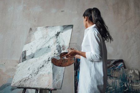 Photo for Young artist beauty swarthy woman in white long shirt bare legs posing against her paintings. Stylish black curly hair sensual african american model with paint palette and brushes at painting studio - Royalty Free Image