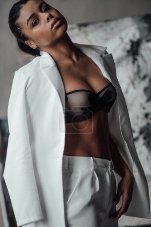 Young adult beauty swarthy woman in formal evening white suit with bra at thoughtful. Stylish black curly hair sensual african american model fashionista posing at studio in fashion pantsuit