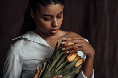 Photo for Young adult beauty swarthy woman in white shirt bare shoulder hugs bouquet of yellow tulips. Stylish black curly hair sensual african american model fashionista posing in studio at spring holidays - Royalty Free Image