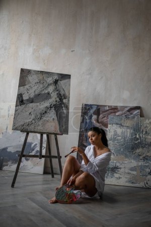 Photo for Young artist beauty swarthy woman in white long shirt bare legs posing against her paintings. Stylish black curly hair sensual african american model with paint palette and brushes at painting studio - Royalty Free Image