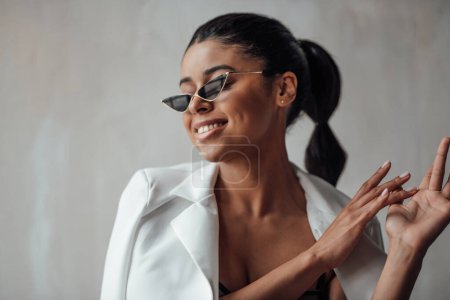 Photo for Young adult beauty swarthy woman in formal evening white suit with bra at cat eye sunglasses. Stylish black curly hair sensual african american model fashionista posing at studio in fashion pantsuit - Royalty Free Image
