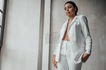 Photo for Young adult beauty swarthy woman in formal evening white suit with bra at thoughtful. Stylish black curly hair sensual african american model fashionista posing at studio in fashion pantsuit - Royalty Free Image