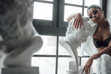 Young beauty swarthy woman in formal evening white suit no shirt hugs greek man bust statue. Stylish black curly hair sensual african american model fashionista posing at studio in fashion pantsuit