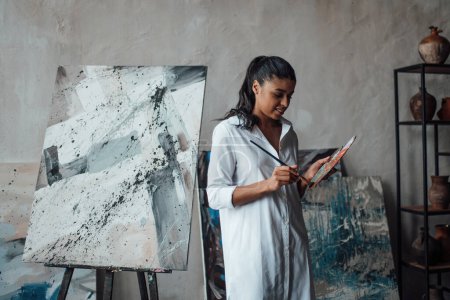 Young artist beauty swarthy woman in white long shirt bare legs posing against her paintings. Stylish black curly hair sensual african american model with paint palette and brushes at painting studio